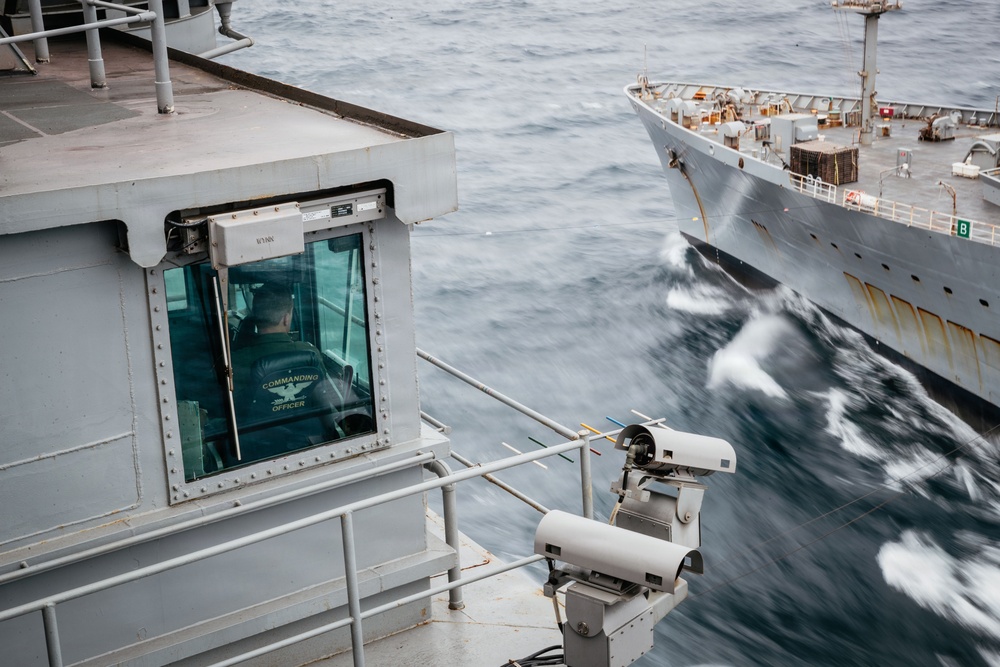 The image shows a view from the deck of a naval vessel, with a person seated in what appears to be a command or observation station labeled 'Commanding Officer.' A second gray military or cargo ship is visible in close proximity on rough seas, suggesting a replenishment-at-sea or close-quarters naval operation. Surveillance or tracking equipment is mounted on the deck railing in the foreground.