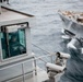 The image shows a view from the deck of a naval vessel, with a person seated in what appears to be a command or observation station labeled 'Commanding Officer.' A second gray military or cargo ship is visible in close proximity on rough seas, suggesting a replenishment-at-sea or close-quarters naval operation. Surveillance or tracking equipment is mounted on the deck railing in the foreground.
