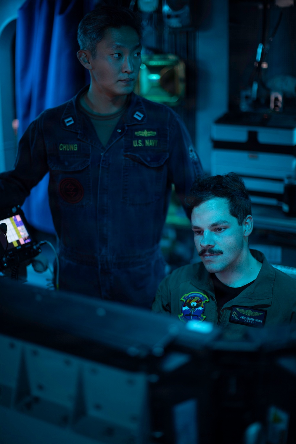 Two U.S. Navy personnel are working together in what appears to be a dimly lit operations or control room aboard a naval vessel. The standing officer, identified by his name tape as 'CHUNG,' wears a U.S. Navy coverall with lieutenant rank insignia and a submarine warfare qualification badge. The seated individual, identified as 'AW02 ANDREW HENKIN,' wears an olive flight suit with a squadron patch and appears to be reviewing information on a monitor.