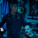 Two U.S. Navy personnel are working together in what appears to be a dimly lit operations or control room aboard a naval vessel. The standing officer, identified by his name tape as 'CHUNG,' wears a U.S. Navy coverall with lieutenant rank insignia and a submarine warfare qualification badge. The seated individual, identified as 'AW02 ANDREW HENKIN,' wears an olive flight suit with a squadron patch and appears to be reviewing information on a monitor.