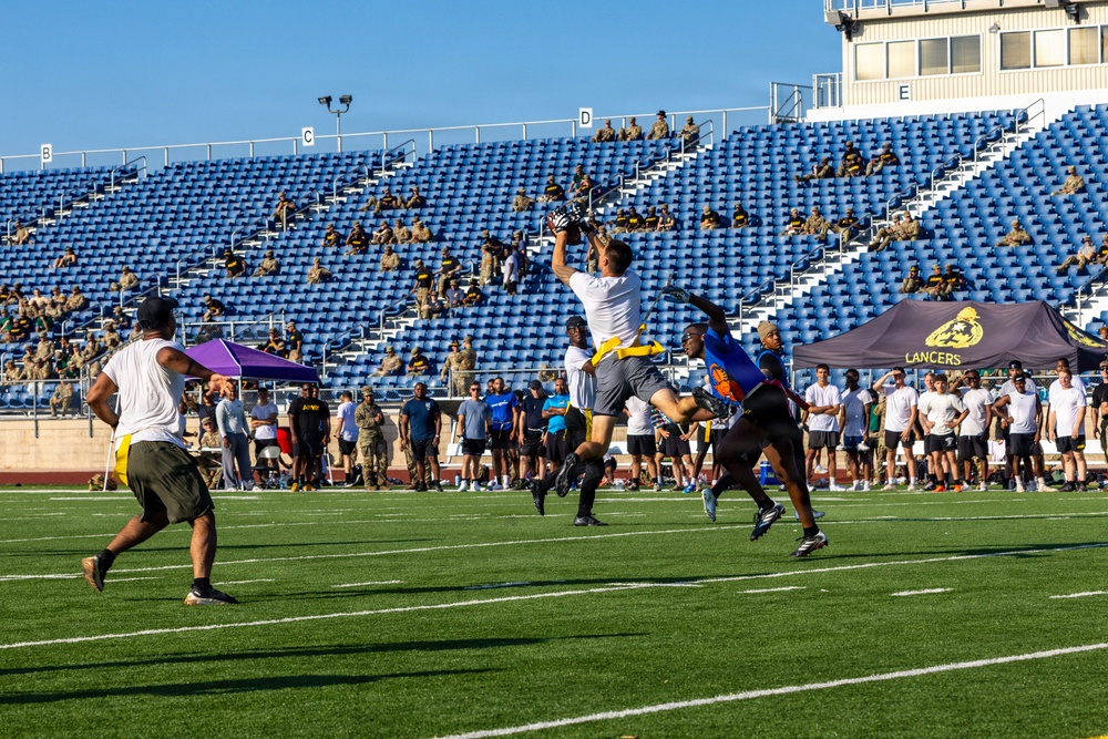 A flag football game is being played on a turf field at a stadium with blue seats. A player in white is leaping to catch or intercept a pass while defenders in dark uniforms contest the play. Spectators, many appearing to be in military uniforms, are seated in the stands and lining the sidelines.