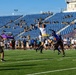 A flag football game is being played on a turf field at a stadium with blue seats. A player in white is leaping to catch or intercept a pass while defenders in dark uniforms contest the play. Spectators, many appearing to be in military uniforms, are seated in the stands and lining the sidelines.