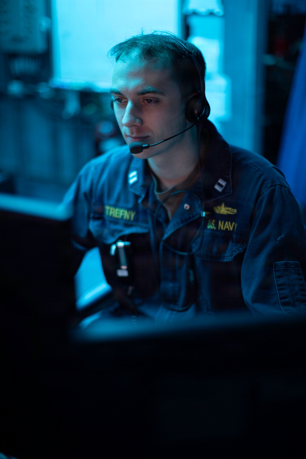 A U.S. Navy personnel member is seated at a workstation in a dimly lit, blue-toned operational environment. The individual is wearing a Navy working uniform with visible name tape reading 'TREFNY' and branch tape reading 'U.S. NAVY', along with a headset with boom microphone. A gold submarine warfare qualification badge is visible on the right chest, suggesting a submarine or naval operations context.