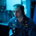 A U.S. Navy personnel member is seated at a workstation in a dimly lit, blue-toned operational environment. The individual is wearing a Navy working uniform with visible name tape reading 'TREFNY' and branch tape reading 'U.S. NAVY', along with a headset with boom microphone. A gold submarine warfare qualification badge is visible on the right chest, suggesting a submarine or naval operations context.