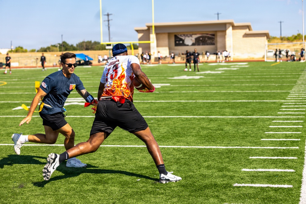 Two players are engaged in a flag football game on a well-maintained artificial turf field. A defender in a navy blue shirt is reaching to pull the flag of an offensive player wearing a white and flame-patterned jersey numbered 13. The game is taking place at what appears to be a military or institutional facility, with a yellow goalpost and a tan building visible in the background.