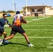 Two players are engaged in a flag football game on a well-maintained artificial turf field. A defender in a navy blue shirt is reaching to pull the flag of an offensive player wearing a white and flame-patterned jersey numbered 13. The game is taking place at what appears to be a military or institutional facility, with a yellow goalpost and a tan building visible in the background.