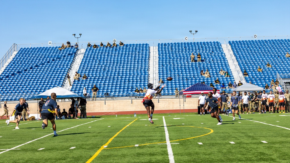 A flag football game is being played on a well-maintained artificial turf field at a stadium with blue seating sections labeled A through D. Military personnel in uniform are visible both in the stands and along the sidelines, suggesting this is a military-organized sporting event. An American flag is displayed prominently along the sideline, reinforcing the military context of the event.