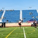 A flag football game is being played on a well-maintained artificial turf field at a stadium with blue seating sections labeled A through D. Military personnel in uniform are visible both in the stands and along the sidelines, suggesting this is a military-organized sporting event. An American flag is displayed prominently along the sideline, reinforcing the military context of the event.
