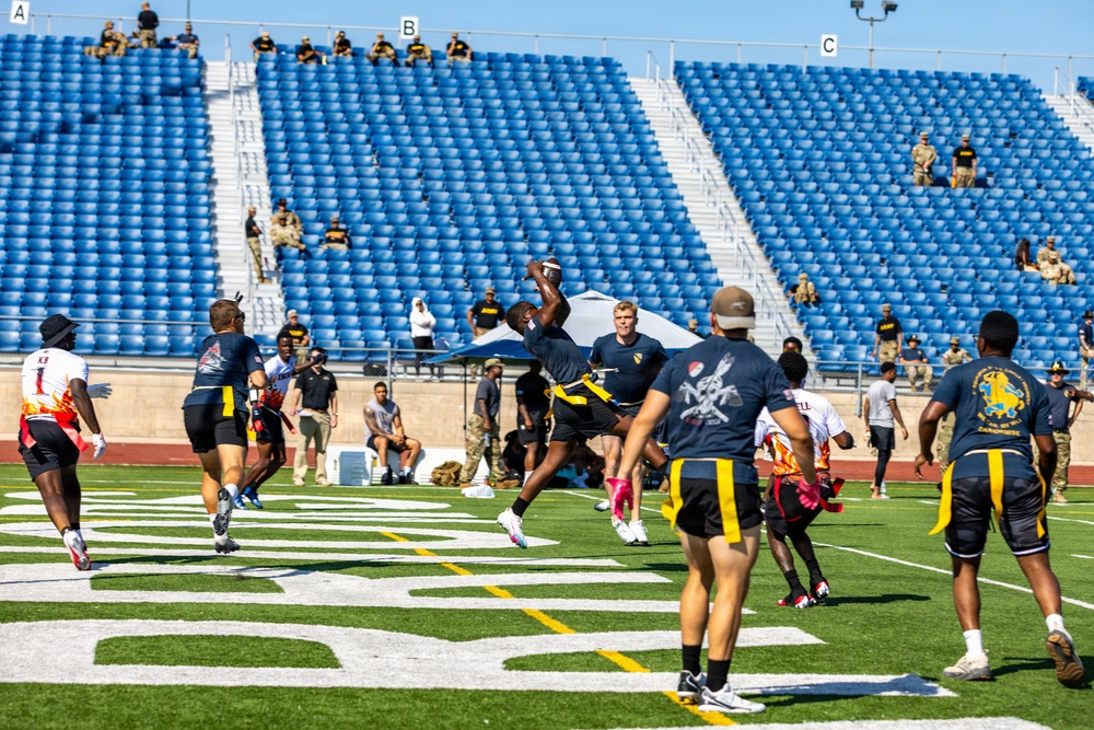A flag football game is being played on a well-maintained turf field inside a stadium with predominantly empty blue seats. Players wearing dark navy and yellow uniforms compete against players in white and red uniforms, with military personnel visible both on the sidelines and in the stands as spectators. The action centers on a player leaping to catch or intercept a football near what appears to be an end zone.
