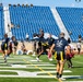 A flag football game is being played on a well-maintained turf field inside a stadium with predominantly empty blue seats. Players wearing dark navy and yellow uniforms compete against players in white and red uniforms, with military personnel visible both on the sidelines and in the stands as spectators. The action centers on a player leaping to catch or intercept a football near what appears to be an end zone.