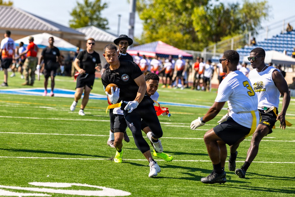 A flag football game is in progress on a well-maintained artificial turf field. A player in black is carrying the football while being pursued by defenders wearing white and black uniforms with yellow flags. The event appears to be a military-affiliated flag football tournament, evidenced by 'ARMY' text visible on players' shorts.