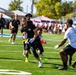 A flag football game is in progress on a well-maintained artificial turf field. A player in black is carrying the football while being pursued by defenders wearing white and black uniforms with yellow flags. The event appears to be a military-affiliated flag football tournament, evidenced by 'ARMY' text visible on players' shorts.