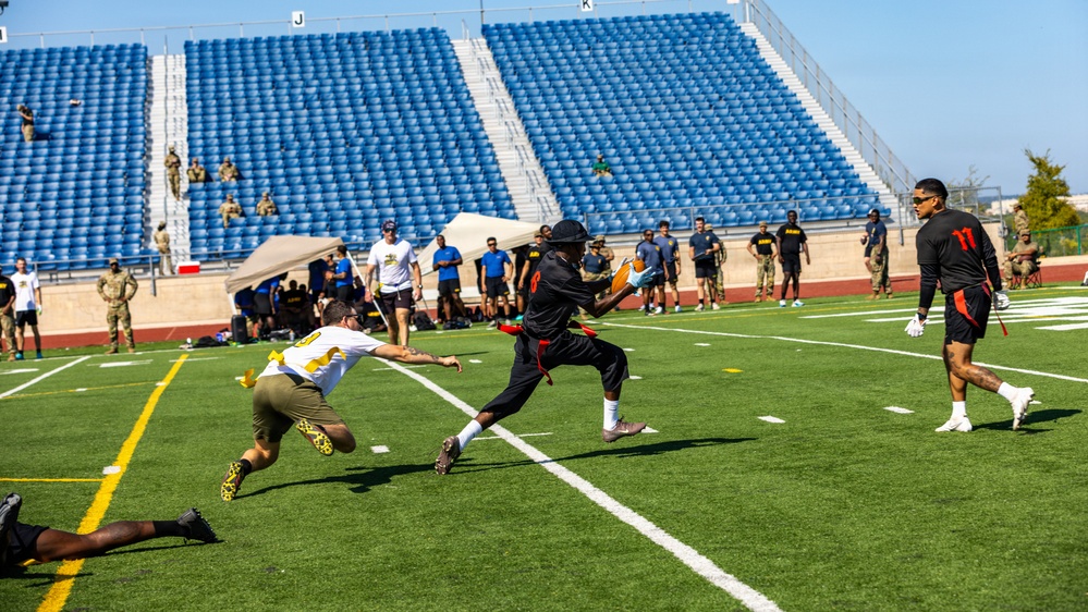 A flag football game is being played on a green turf field in a stadium with blue empty bleachers in the background. A player in black is carrying the ball while being pursued by a defender in white and yellow who is lunging to pull a flag. Spectators and what appear to be military personnel in uniform line the sidelines.