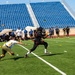 A flag football game is being played on a green turf field in a stadium with blue empty bleachers in the background. A player in black is carrying the ball while being pursued by a defender in white and yellow who is lunging to pull a flag. Spectators and what appear to be military personnel in uniform line the sidelines.