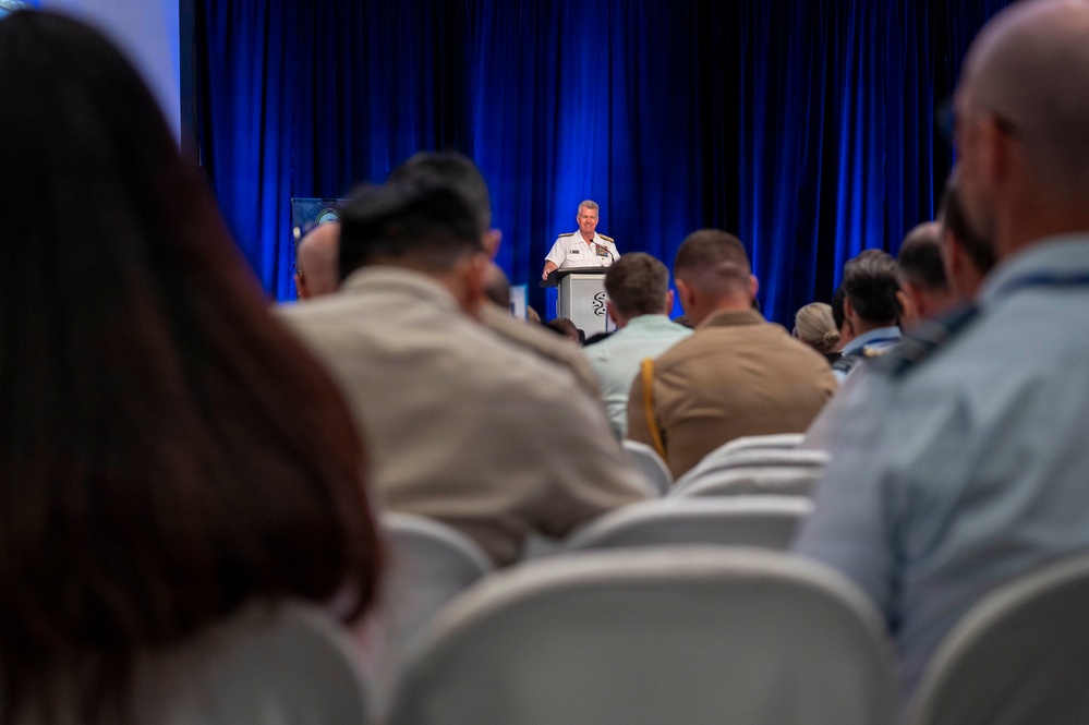 A military officer in white dress uniform is delivering a speech at a podium during a formal conference or symposium. The audience consists of a mixed group of military personnel in various uniforms and civilians seated in rows of white chairs. The setting features a blue curtained backdrop and a branded lectern on a stage.