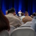 A military officer in white dress uniform is delivering a speech at a podium during a formal conference or symposium. The audience consists of a mixed group of military personnel in various uniforms and civilians seated in rows of white chairs. The setting features a blue curtained backdrop and a branded lectern on a stage.