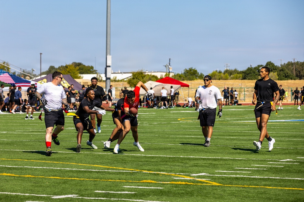 A flag football game is being played on a well-maintained artificial turf field under clear blue skies. A player in a red jersey is carrying the football while being pursued by defenders wearing black and white attire. The setting appears to be an organized recreational or military event, evidenced by tents, an American flag, and a large crowd of spectators in the background.