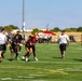 A flag football game is being played on a well-maintained artificial turf field under clear blue skies. A player in a red jersey is carrying the football while being pursued by defenders wearing black and white attire. The setting appears to be an organized recreational or military event, evidenced by tents, an American flag, and a large crowd of spectators in the background.