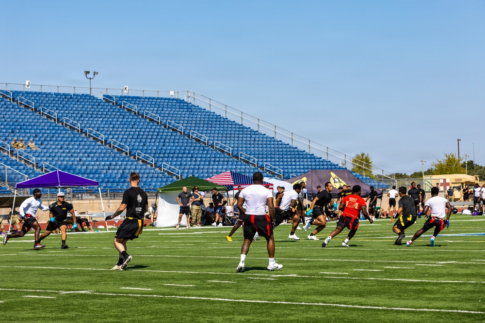 A flag football game is being played on a green turf field at a stadium with blue bleacher seating. Players wearing black and gold shorts with various colored shirts are actively engaged in a play, with sideline tents and an American flag visible in the background. The setting appears to be a military installation based on visible Army-related banners and camouflage-uniformed personnel.