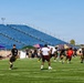 A flag football game is being played on a green turf field at a stadium with blue bleacher seating. Players wearing black and gold shorts with various colored shirts are actively engaged in a play, with sideline tents and an American flag visible in the background. The setting appears to be a military installation based on visible Army-related banners and camouflage-uniformed personnel.