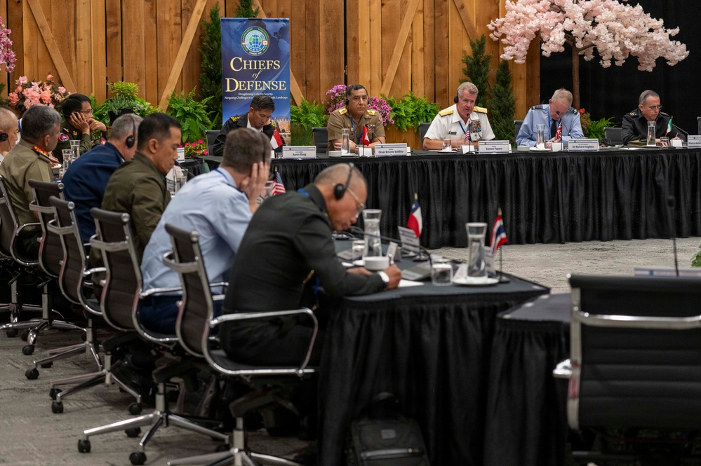 A multinational military conference titled 'Chiefs of Defense' is taking place in a large venue decorated with floral arrangements and wooden panel backdrops. Senior military officers in various national uniforms are seated at a large conference table, with name placards visible in front of them. The event appears to be a high-level defense summit focused on collective security, as indicated by the banner subtitle referencing 'Strengthening Collective Security.'