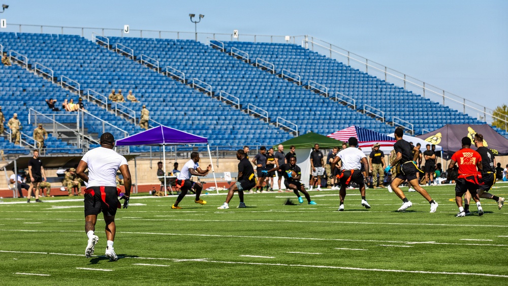 An outdoor flag football or 7-on-7 style game is taking place on a well-maintained artificial turf field inside a stadium with blue empty seats. Players wearing black-and-gold uniforms compete against players in white-and-red uniforms, with military personnel in camouflage uniforms visible in the stands and along the sidelines. Several event tents, including one displaying an American flag, are set up along the sideline, suggesting a military-sponsored or military-affiliated sporting event.