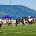 An outdoor flag football or 7-on-7 style game is taking place on a well-maintained artificial turf field inside a stadium with blue empty seats. Players wearing black-and-gold uniforms compete against players in white-and-red uniforms, with military personnel in camouflage uniforms visible in the stands and along the sidelines. Several event tents, including one displaying an American flag, are set up along the sideline, suggesting a military-sponsored or military-affiliated sporting event.