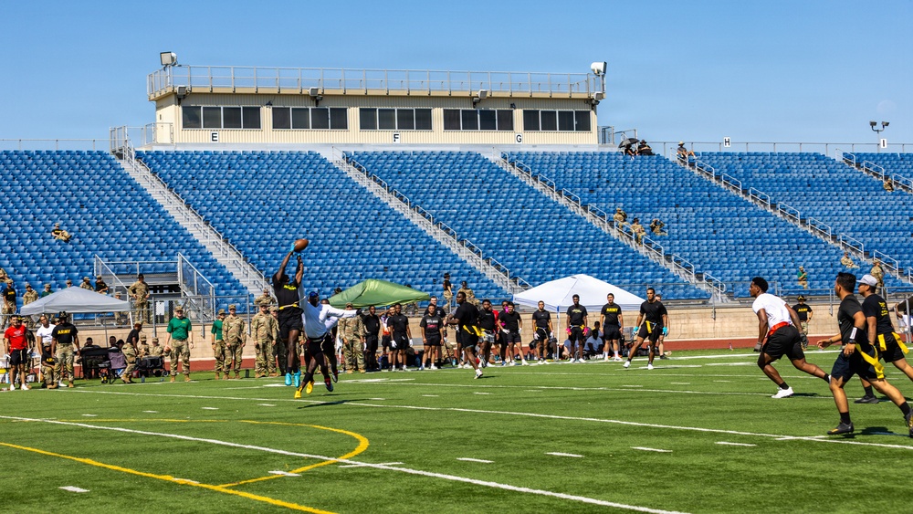 A flag football game is being played on a green turf field at a stadium with blue empty bleachers in the background. Players wearing black and yellow uniforms, consistent with U.S. Army branding, are actively competing, with one player leaping to catch a football. Military personnel in camouflage uniforms are visible along the sidelines as spectators.