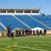 A flag football game is being played on a green turf field at a stadium with blue empty bleachers in the background. Players wearing black and yellow uniforms, consistent with U.S. Army branding, are actively competing, with one player leaping to catch a football. Military personnel in camouflage uniforms are visible along the sidelines as spectators.
