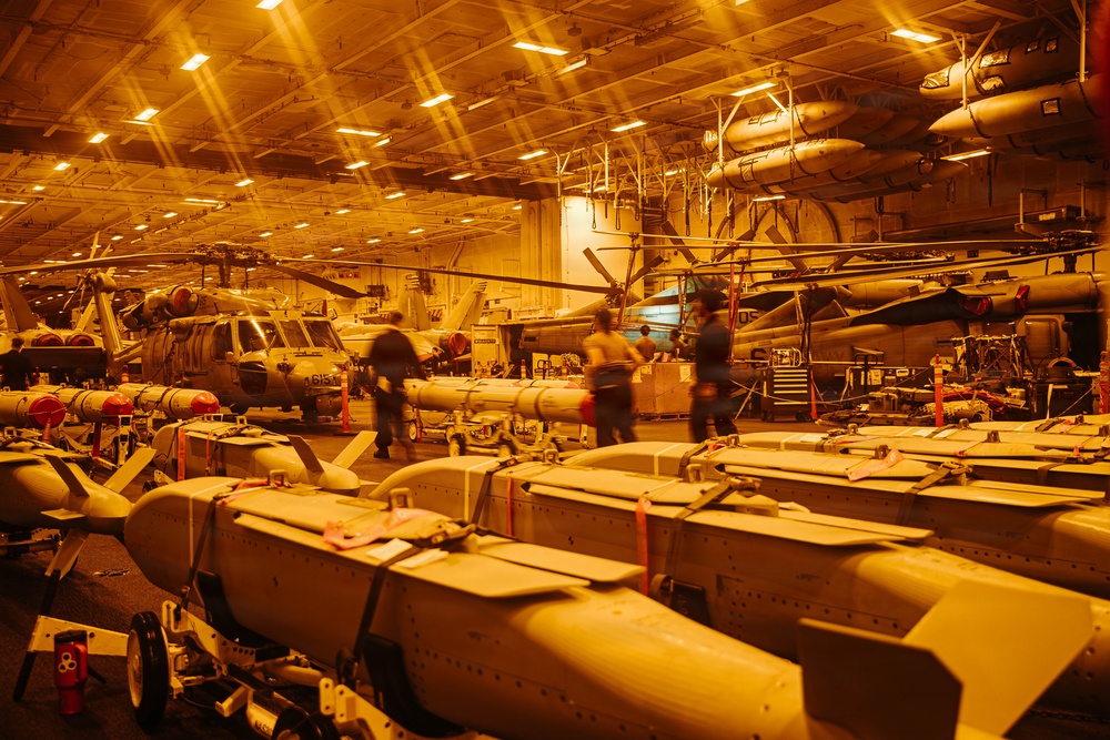 The image shows the interior of a naval vessel's hangar bay, likely an aircraft carrier, illuminated by warm orange overhead lighting. Multiple military helicopters are stored with folded rotor blades alongside rows of ordnance/munitions staged on carts in the foreground. Several personnel are visible moving through the space, appearing motion-blurred due to a longer camera exposure.