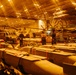 The image shows the interior of a naval vessel's hangar bay, likely an aircraft carrier, illuminated by warm orange overhead lighting. Multiple military helicopters are stored with folded rotor blades alongside rows of ordnance/munitions staged on carts in the foreground. Several personnel are visible moving through the space, appearing motion-blurred due to a longer camera exposure.