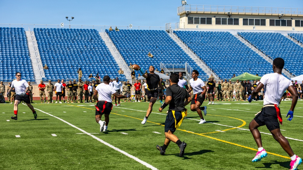 A flag football game is being played on a well-maintained artificial turf field inside a stadium with blue seats. Players wearing two distinct uniforms — black/gold with flag belts and white/red with flag belts — are actively competing, with one player leaping to catch a football. A large crowd of spectators, many appearing to be in military uniforms (Army), lines the sidelines and background.