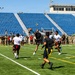 A flag football game is being played on a well-maintained artificial turf field inside a stadium with blue seats. Players wearing two distinct uniforms — black/gold with flag belts and white/red with flag belts — are actively competing, with one player leaping to catch a football. A large crowd of spectators, many appearing to be in military uniforms (Army), lines the sidelines and background.