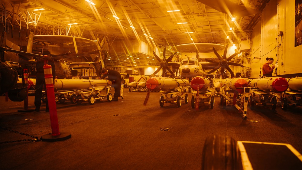 The image shows the interior of a naval aircraft carrier hangar bay illuminated by warm orange/amber lighting. Multiple aircraft are stored inside, including what appears to be an E-2 Hawkeye (marked '604') with distinctive multi-blade propellers. Numerous ordnance/munitions on wheeled carts are staged in rows across the hangar deck, with personnel visible working among the equipment.