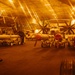 The image shows the interior of a naval aircraft carrier hangar bay illuminated by warm orange/amber lighting. Multiple aircraft are stored inside, including what appears to be an E-2 Hawkeye (marked '604') with distinctive multi-blade propellers. Numerous ordnance/munitions on wheeled carts are staged in rows across the hangar deck, with personnel visible working among the equipment.