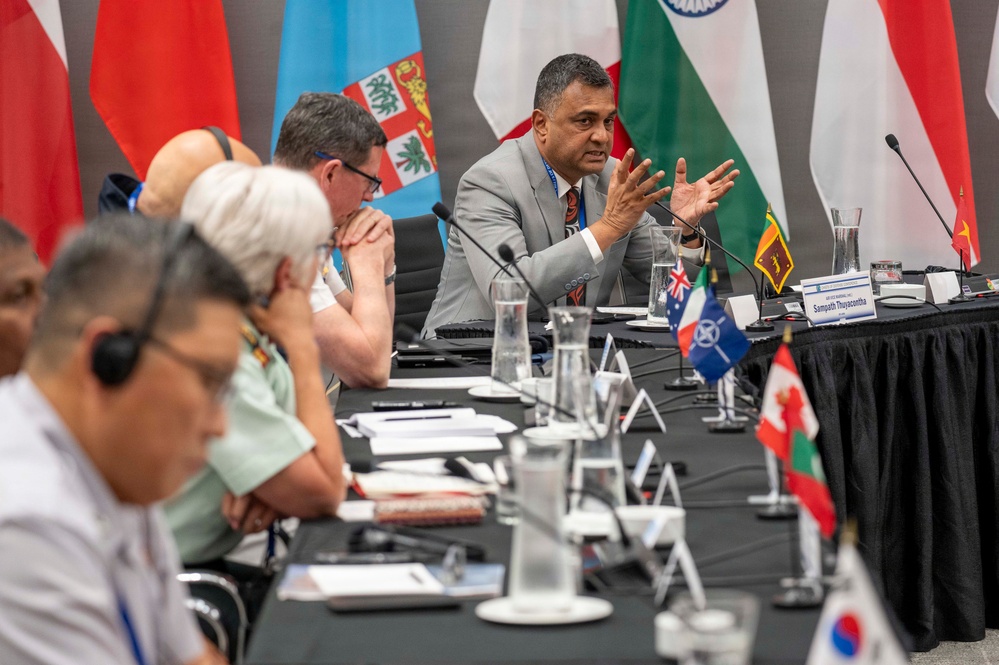 A multinational conference or diplomatic meeting is taking place at a long conference table. A man in a gray suit, identified by a nameplate as 'Sampath Thuyacontha', is actively speaking with gesturing hands. Multiple national flags are displayed in the background, and small desk flags representing various countries are placed along the table.