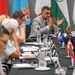 A multinational conference or diplomatic meeting is taking place at a long conference table. A man in a gray suit, identified by a nameplate as 'Sampath Thuyacontha', is actively speaking with gesturing hands. Multiple national flags are displayed in the background, and small desk flags representing various countries are placed along the table.