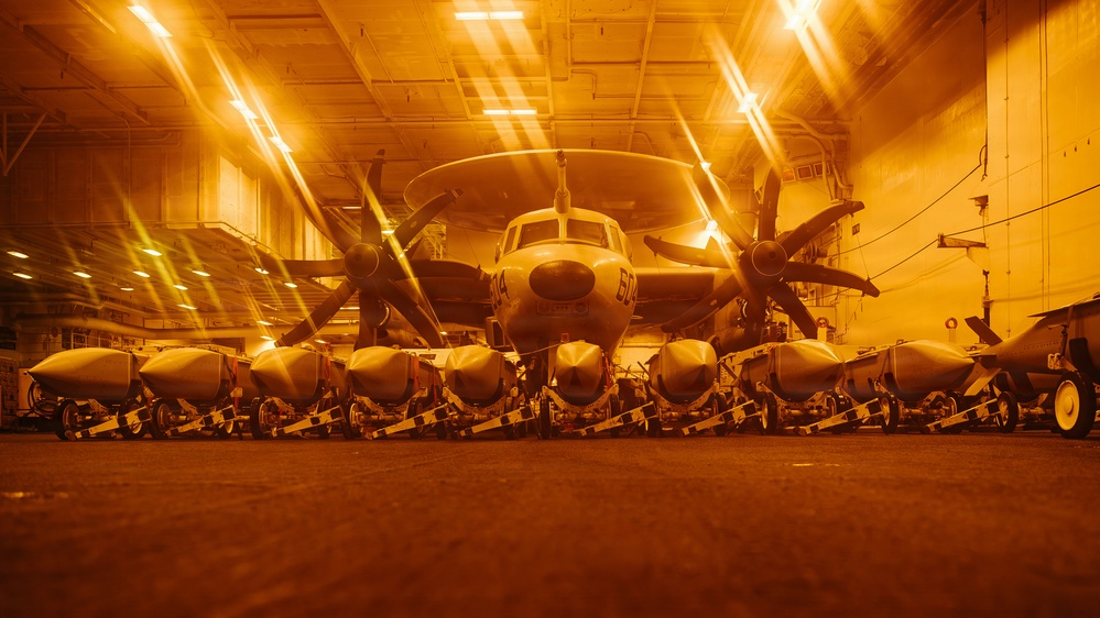 A military aircraft, identifiable as a Grumman E-2 Hawkeye (marked '604'), is stored in what appears to be a naval vessel hangar bay under amber/orange lighting. In the foreground, a row of external fuel tanks or ordnance pods are lined up on wheeled carts. The dramatic low-angle perspective and warm lighting create a striking visual composition of the hangar interior.