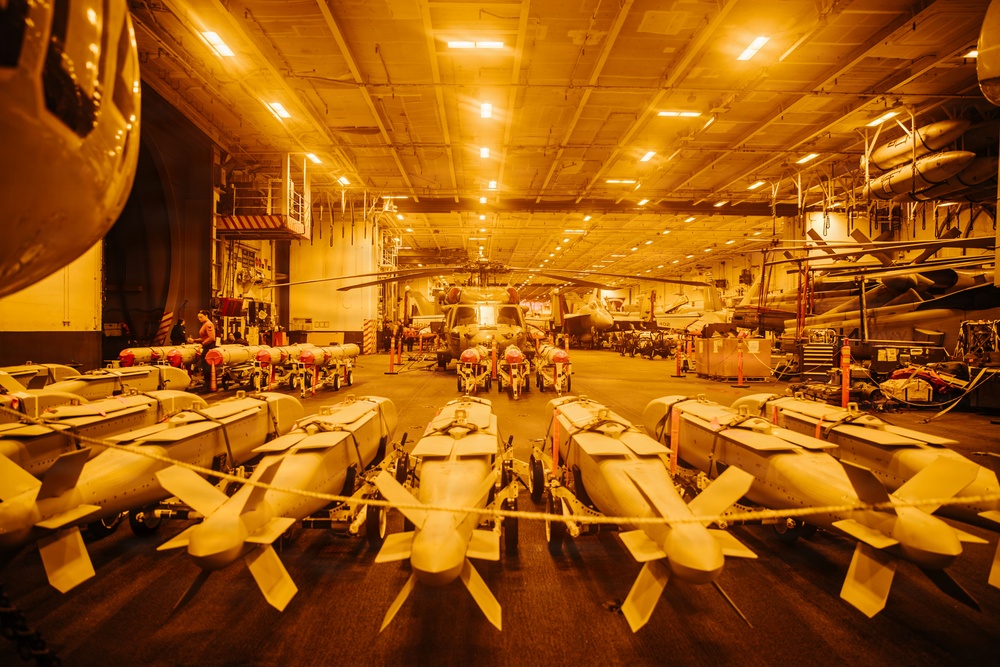 The image shows the interior of a naval vessel's hangar bay, likely an aircraft carrier or amphibious assault ship, illuminated by warm amber/orange lighting. The foreground is dominated by rows of what appear to be aerial munitions (bombs or missiles) secured on wheeled carts. In the background, several military rotary-wing and fixed-wing aircraft are visible, along with maintenance personnel.