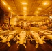 The image shows the interior of a naval vessel's hangar bay, likely an aircraft carrier or amphibious assault ship, illuminated by warm amber/orange lighting. The foreground is dominated by rows of what appear to be aerial munitions (bombs or missiles) secured on wheeled carts. In the background, several military rotary-wing and fixed-wing aircraft are visible, along with maintenance personnel.