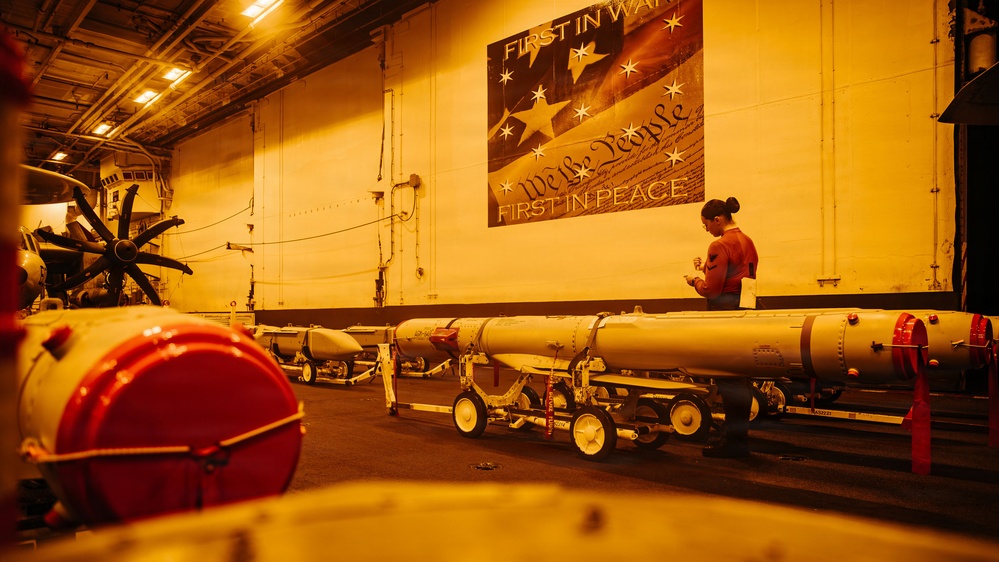 The image shows the interior of what appears to be a naval vessel's hangar bay, likely an aircraft carrier, with multiple large munitions or torpedoes staged on wheeled carts. A single person stands near the wall reviewing documents or a clipboard. A patriotic mural reading 'First in War... We the People... First in Peace' is prominently displayed on the wall.