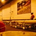 The image shows the interior of what appears to be a naval vessel's hangar bay, likely an aircraft carrier, with multiple large munitions or torpedoes staged on wheeled carts. A single person stands near the wall reviewing documents or a clipboard. A patriotic mural reading 'First in War... We the People... First in Peace' is prominently displayed on the wall.