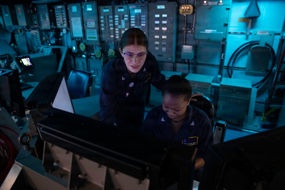 Two female military personnel in navy blue uniforms are working at a control station aboard what appears to be a naval vessel. One sailor is seated at a console while the other stands behind her, both focused on equipment in front of them. The environment is a dimly lit operations or bridge room with extensive control panels and instrumentation visible in the background.