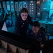 Two female military personnel in navy blue uniforms are working at a control station aboard what appears to be a naval vessel. One sailor is seated at a console while the other stands behind her, both focused on equipment in front of them. The environment is a dimly lit operations or bridge room with extensive control panels and instrumentation visible in the background.