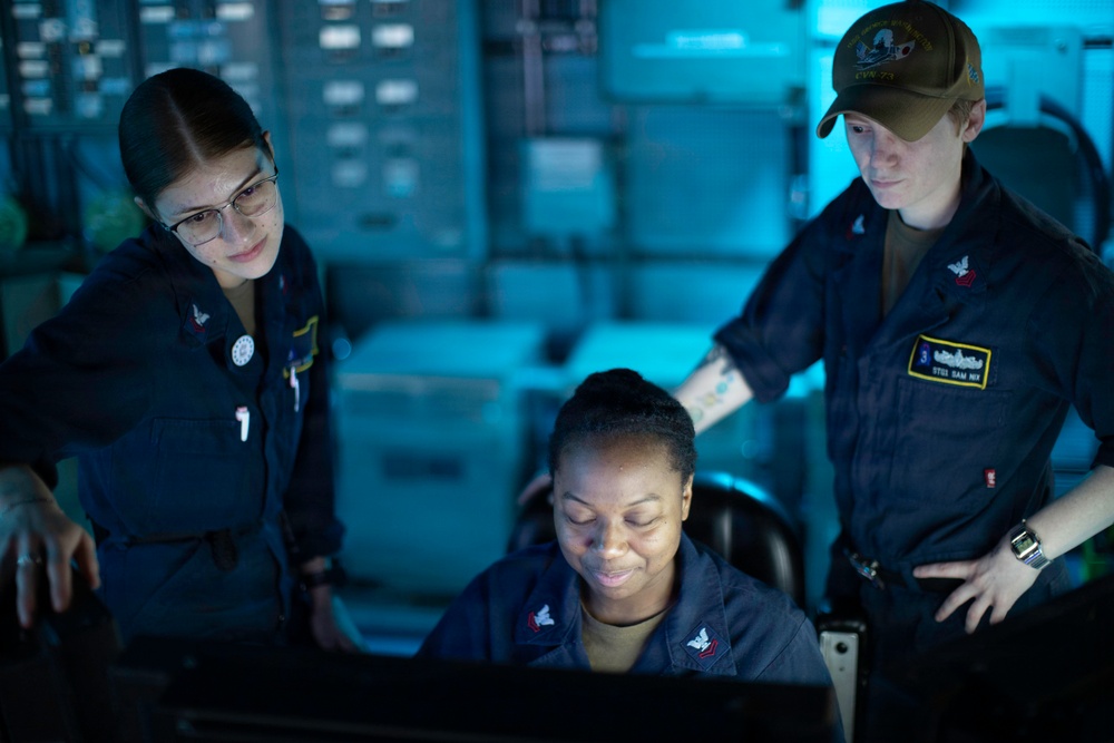 Three U.S. Navy personnel in navy blue coverall uniforms are gathered around a computer workstation in what appears to be a shipboard operations or combat information center. The scene is illuminated primarily by blue ambient lighting and the glow of a monitor screen. Two personnel are standing and observing while one is seated at the workstation.