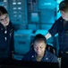 Three U.S. Navy personnel in navy blue coverall uniforms are gathered around a computer workstation in what appears to be a shipboard operations or combat information center. The scene is illuminated primarily by blue ambient lighting and the glow of a monitor screen. Two personnel are standing and observing while one is seated at the workstation.