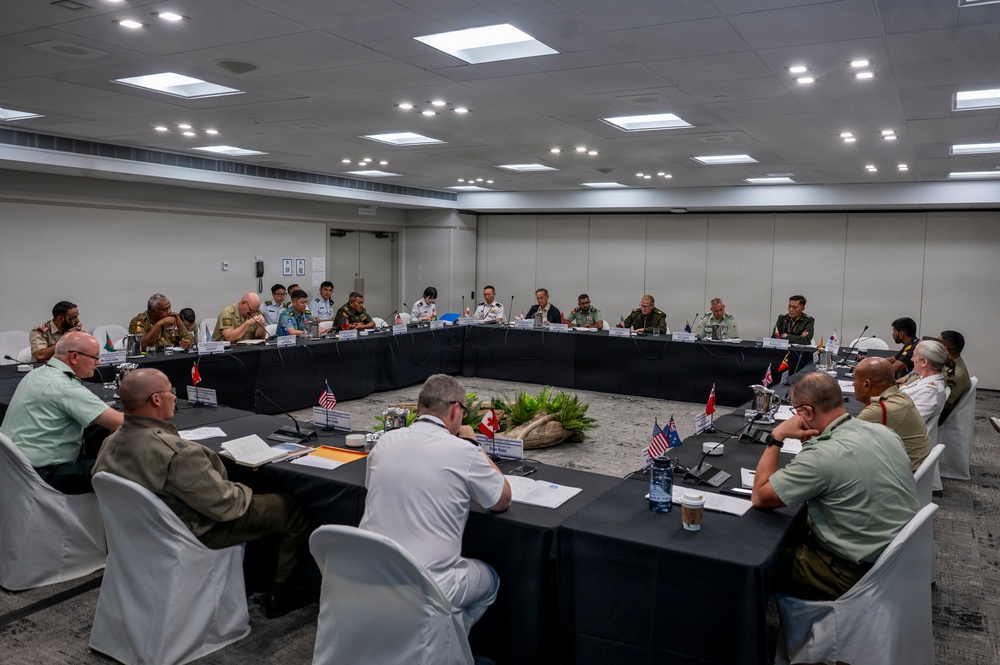 A multinational military conference is taking place in a modern conference room, with uniformed personnel from multiple nations seated around a large U-shaped table arrangement. Small national flags are visible on the table, indicating representation from numerous countries. The setting appears to be a formal high-level military coordination or planning meeting.