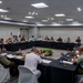 A multinational military conference is taking place in a modern conference room, with uniformed personnel from multiple nations seated around a large U-shaped table arrangement. Small national flags are visible on the table, indicating representation from numerous countries. The setting appears to be a formal high-level military coordination or planning meeting.