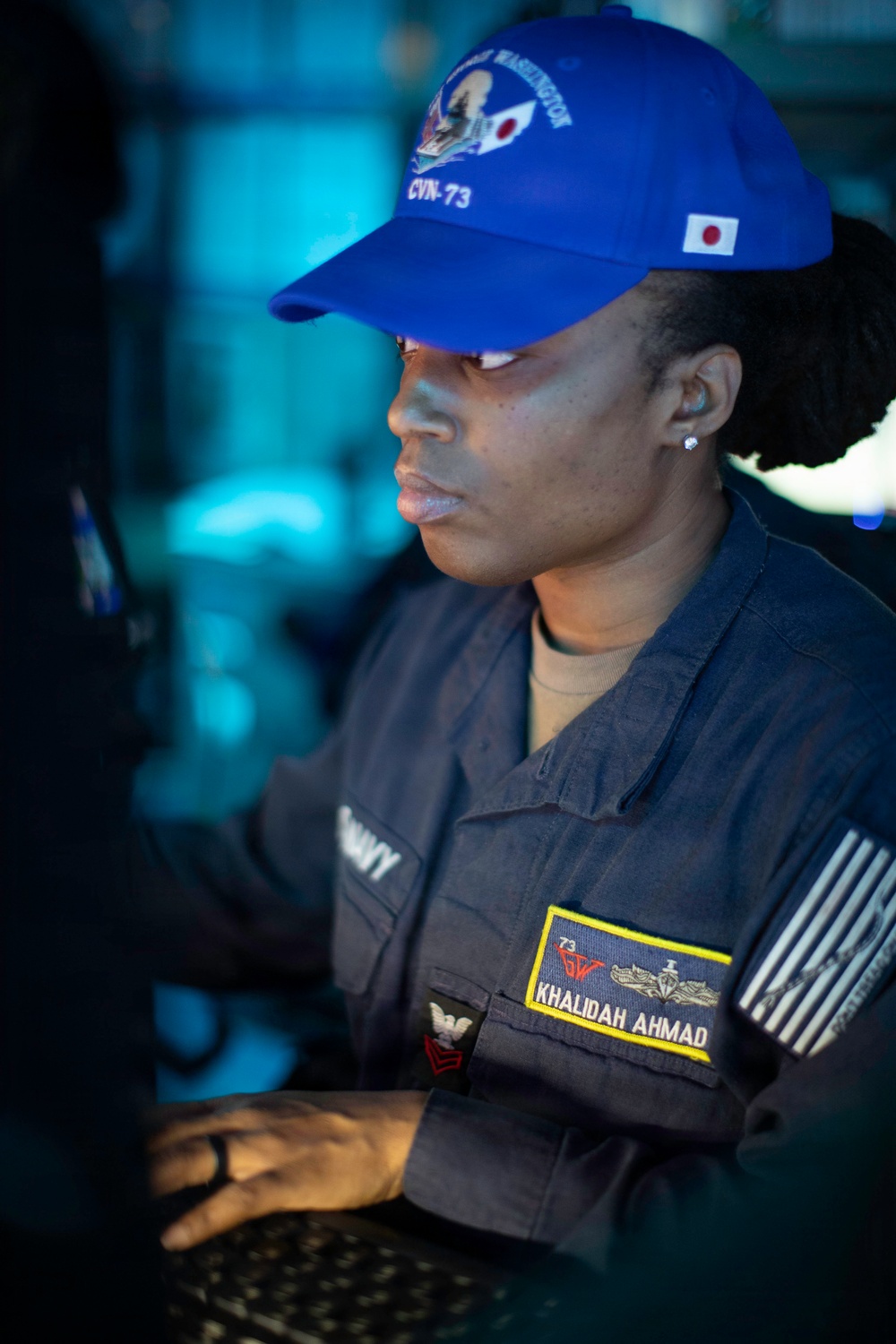 A U.S. Navy sailor named Khalidah Ahmad is shown working at what appears to be a computer terminal or workstation. She is wearing a dark Navy working uniform and a blue baseball cap bearing the designation 'CVN-73' (USS George Washington) with a Japanese flag patch. The image is taken in a dimly lit shipboard environment with blue ambient lighting in the background.