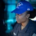 A U.S. Navy sailor named Khalidah Ahmad is shown working at what appears to be a computer terminal or workstation. She is wearing a dark Navy working uniform and a blue baseball cap bearing the designation 'CVN-73' (USS George Washington) with a Japanese flag patch. The image is taken in a dimly lit shipboard environment with blue ambient lighting in the background.